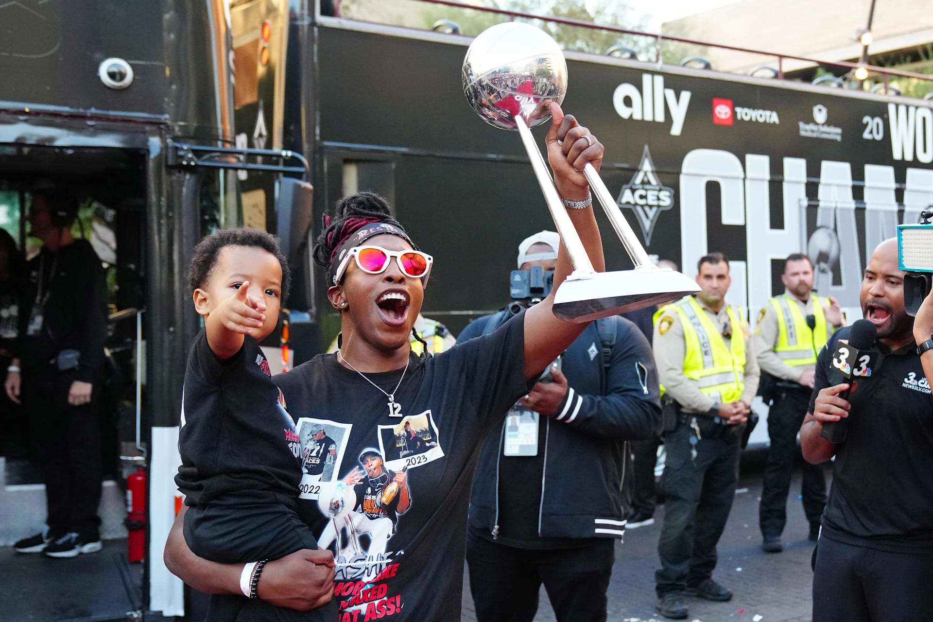 A woman wearing sunglasses celebrates while holding up a championship trophy and carrying a young child, surrounded by a crowd and media in a parade setting with a team bus in the background.