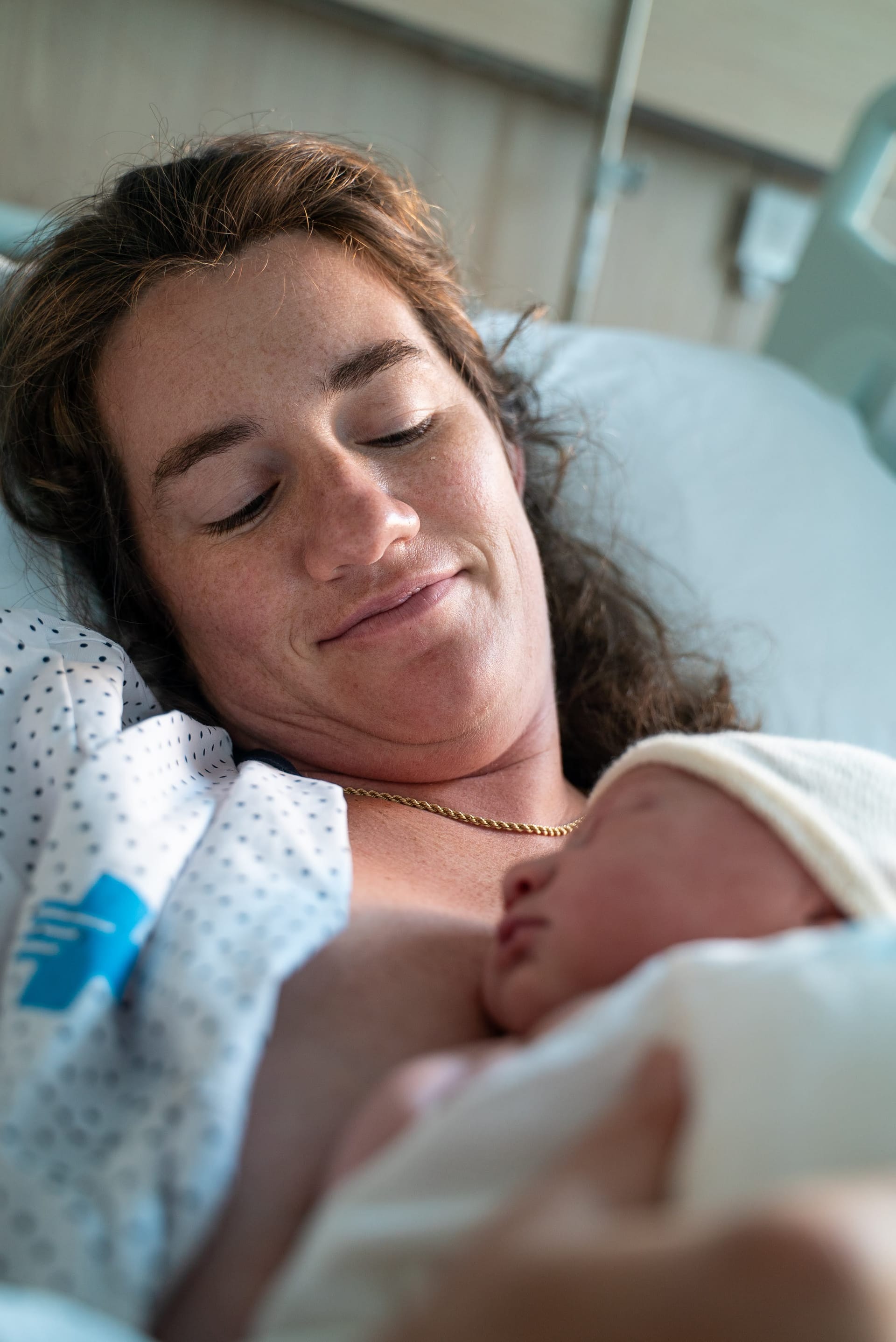 A mother lies in a hospital bed holding her newborn baby against her chest shortly after birth. The baby is wrapped in a white blanket and knit cap while the mother looks down with a soft smile in a quiet hospital room.