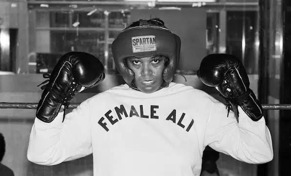 Boxer Jackie Tonawanda holds up gloved fists in a shirt that reads "Female Ali"